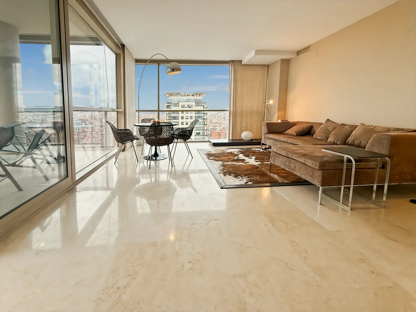 Bright living room with a brown sofa, round table with black chairs, and a cowhide rug. Large windows offer a city view.