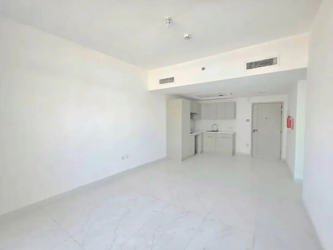Bright, empty studio apartment with white walls, marble floors, and a small kitchen area with light gray cabinets. A door and fire extinguisher are visible.