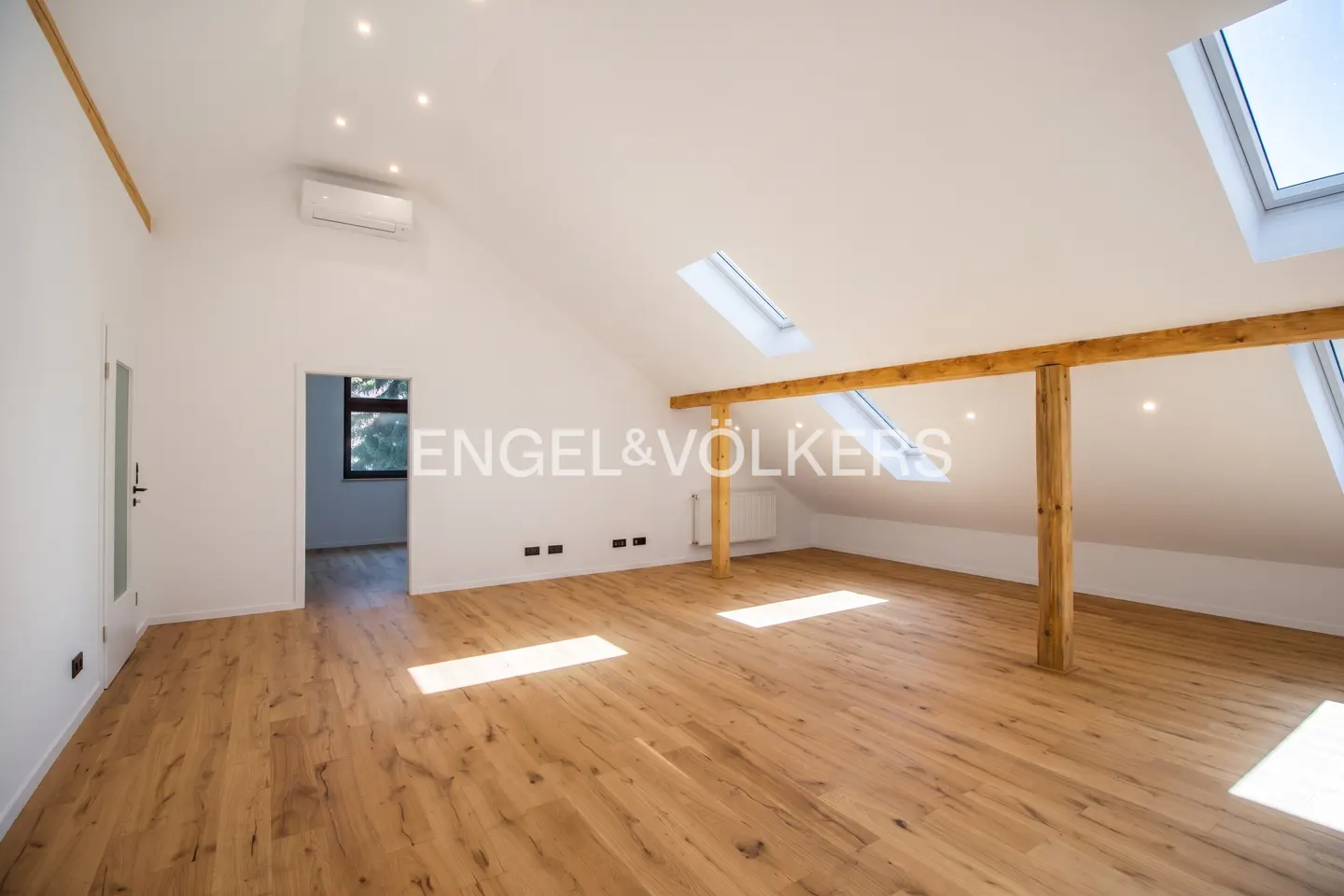 Bright, empty attic room with wood floors, white walls, and exposed beams. Skylights let in natural light. Doorway to another room is visible.