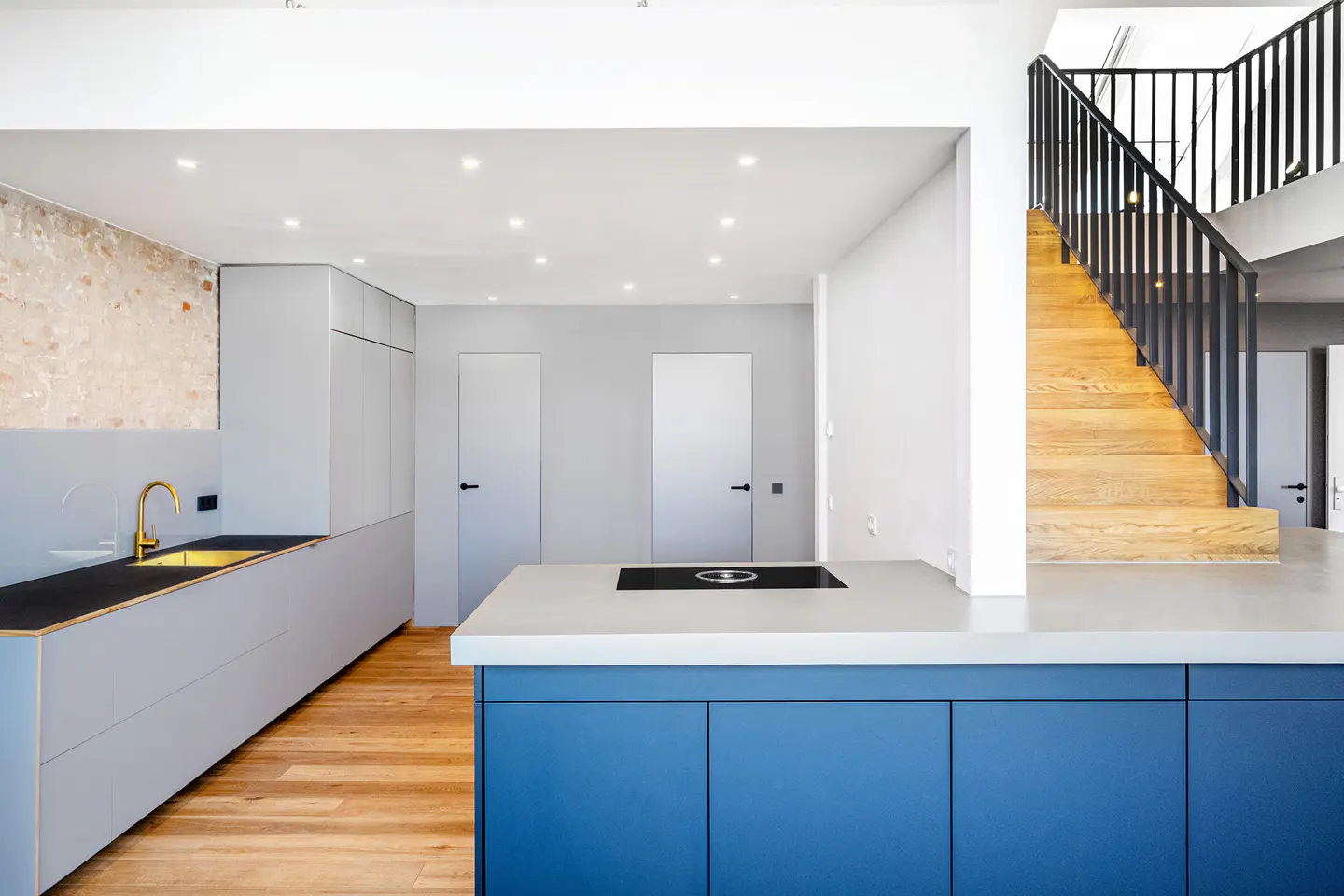 Modern kitchen with blue island, gray cabinets, and gold faucet. Wooden stairs with black railing lead to the second floor.