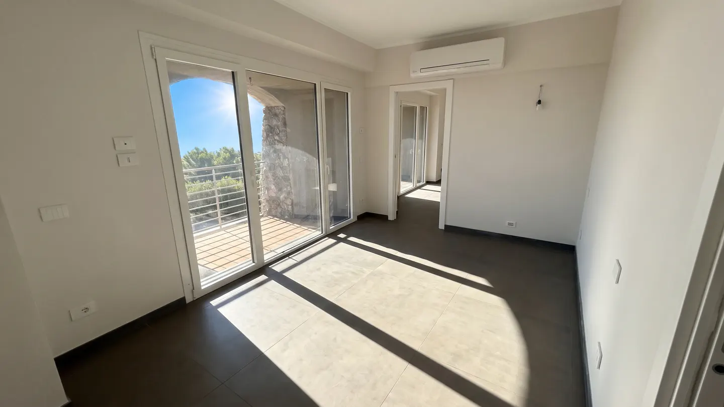 Bright, empty room with white walls, dark tile floor, and sliding glass doors to a balcony with a view of trees and blue sky.