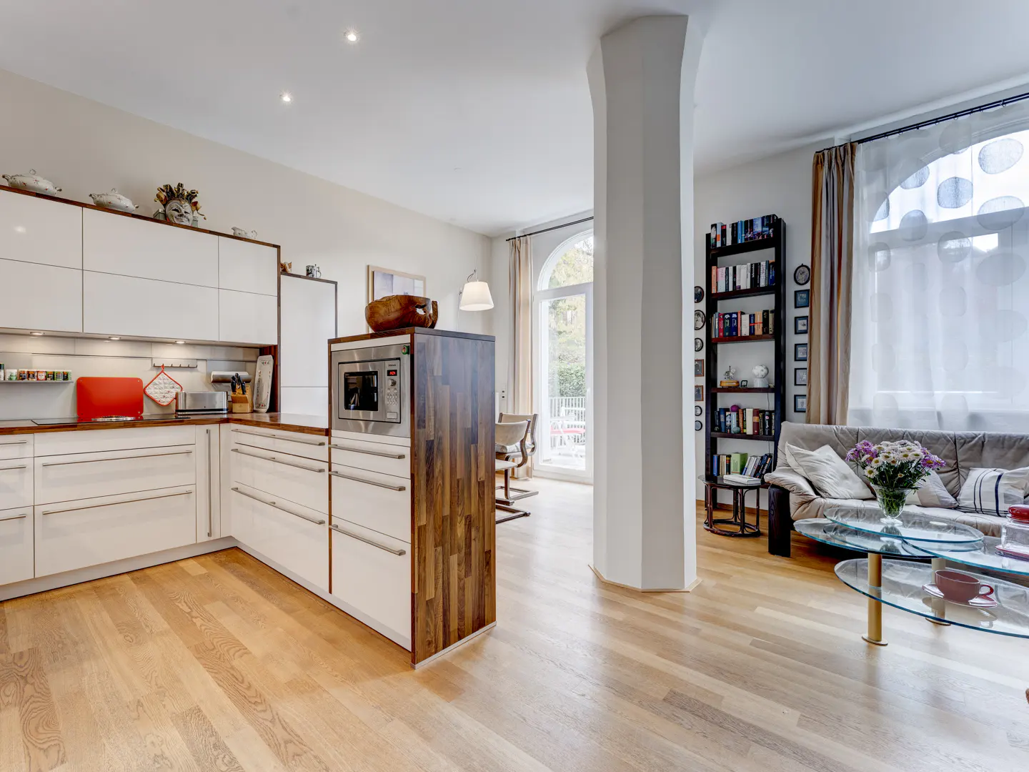 Open-concept living space with white kitchen cabinets, wood floors, and a large white pillar. A living room with a couch and bookshelf is visible.