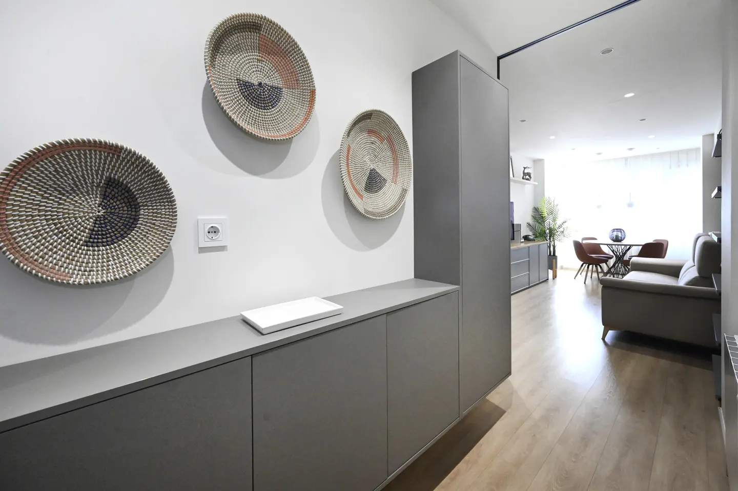 Modern apartment hallway with gray cabinets, decorative woven baskets on the white wall, and a glimpse into the living room.