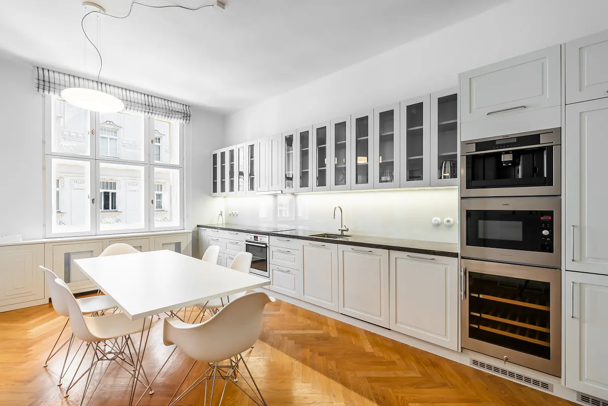 Bright, modern kitchen with white cabinets, stainless steel appliances, and a large window. A white table and chairs sit on a herringbone wood floor.