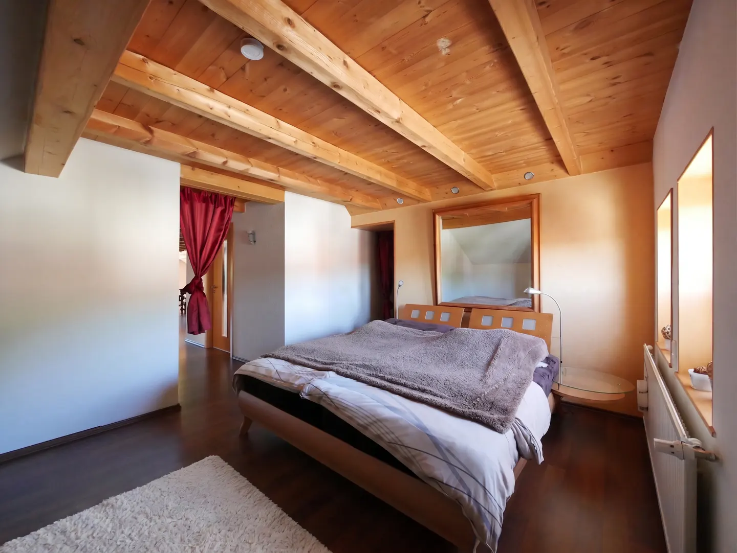 Bedroom with wood ceiling, beams, and floor. A bed with a brown blanket is centered. Red curtains hang in the doorway.