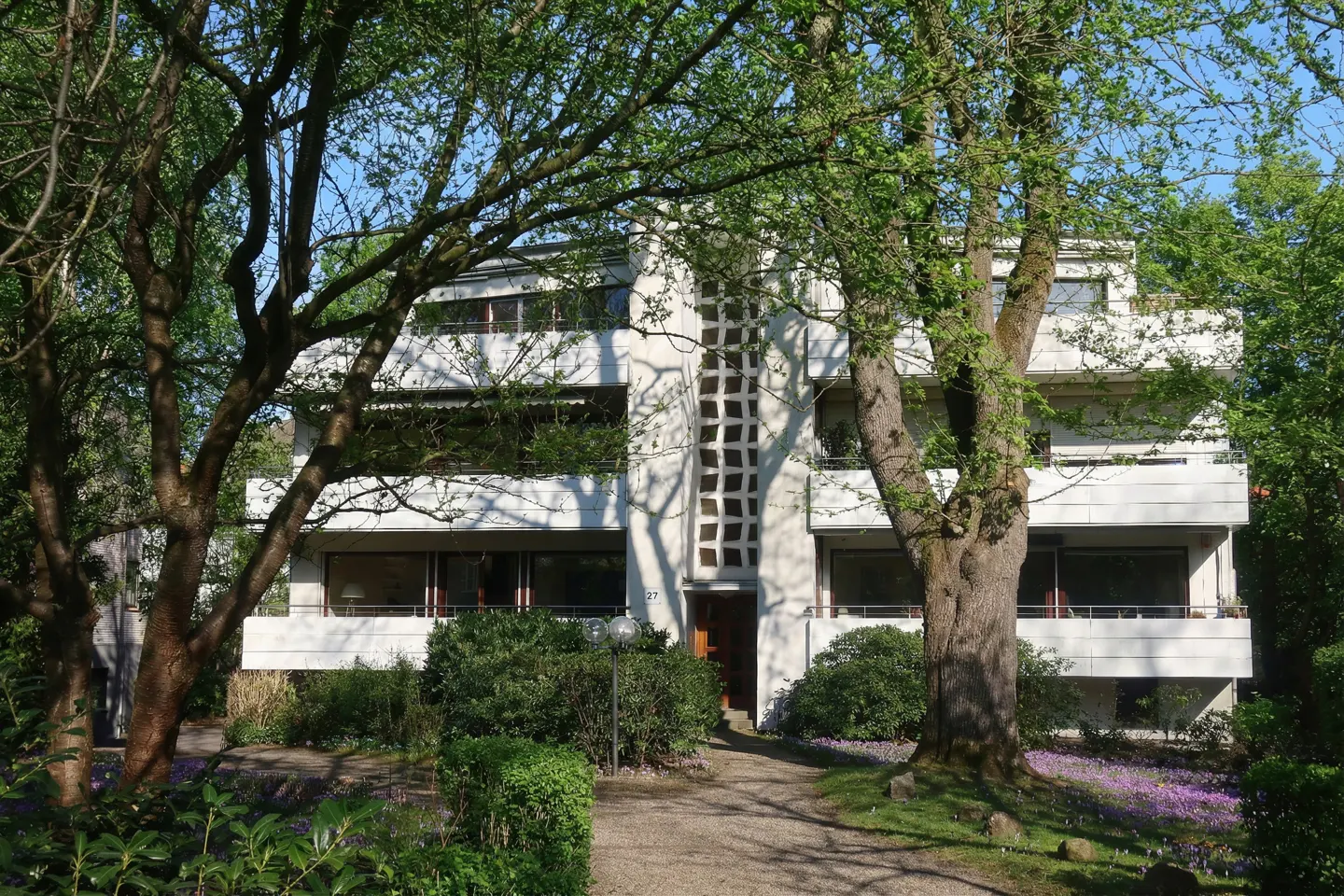 Modern white apartment building with balconies, framed by green trees and foliage. A pathway leads to the entrance.