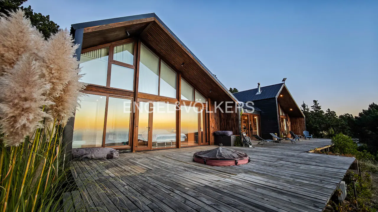Exterior view of a modern wood cabin with large windows and a wooden deck at sunset. Tall pampas grass in the foreground.