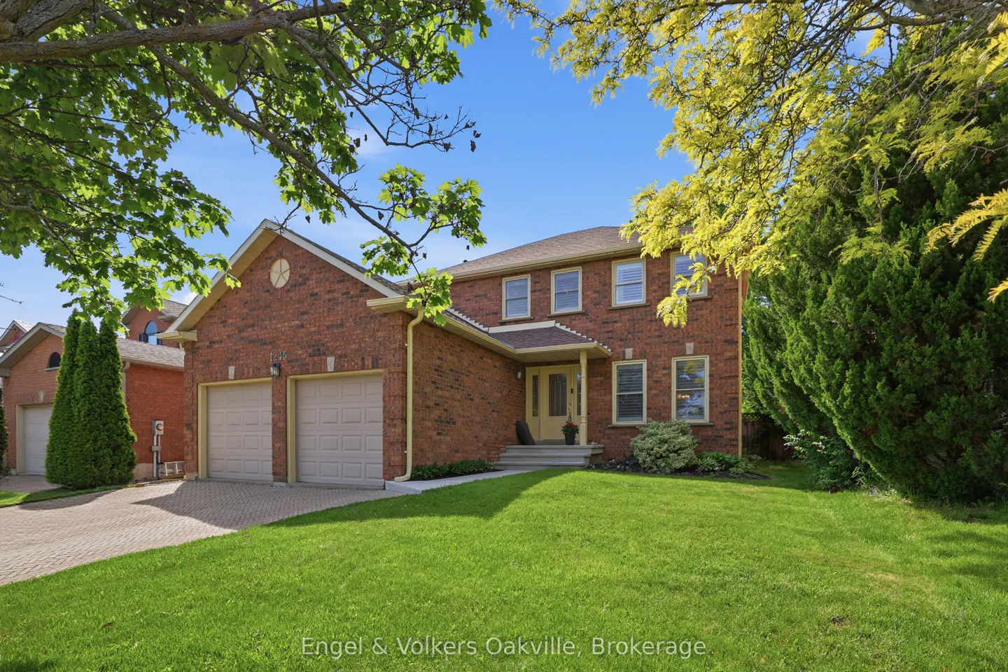 Two-story red brick suburban home with a double garage, green lawn, and mature trees under a clear blue sky.