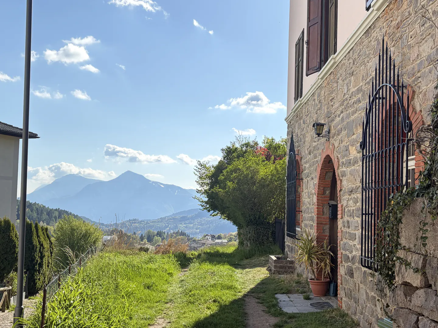 Stone house with arched windows and iron bars overlooks a green hillside and distant mountains under a blue sky.