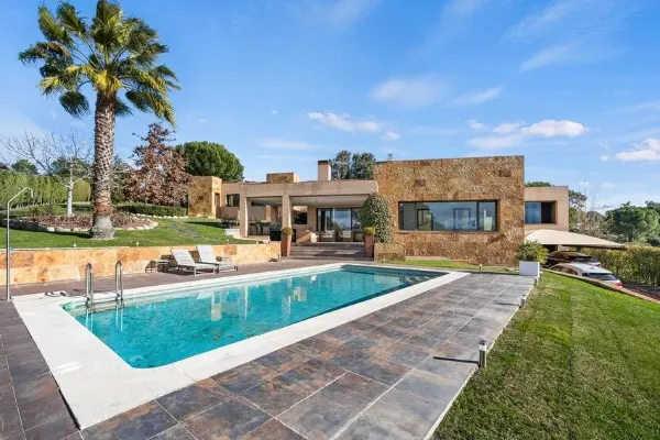 Luxury home exterior with a blue swimming pool, stone patio, and palm tree under a blue sky.