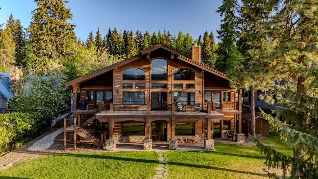 Two-story log cabin with large windows, a stone chimney, and a covered porch. Green lawn and trees surround the house.