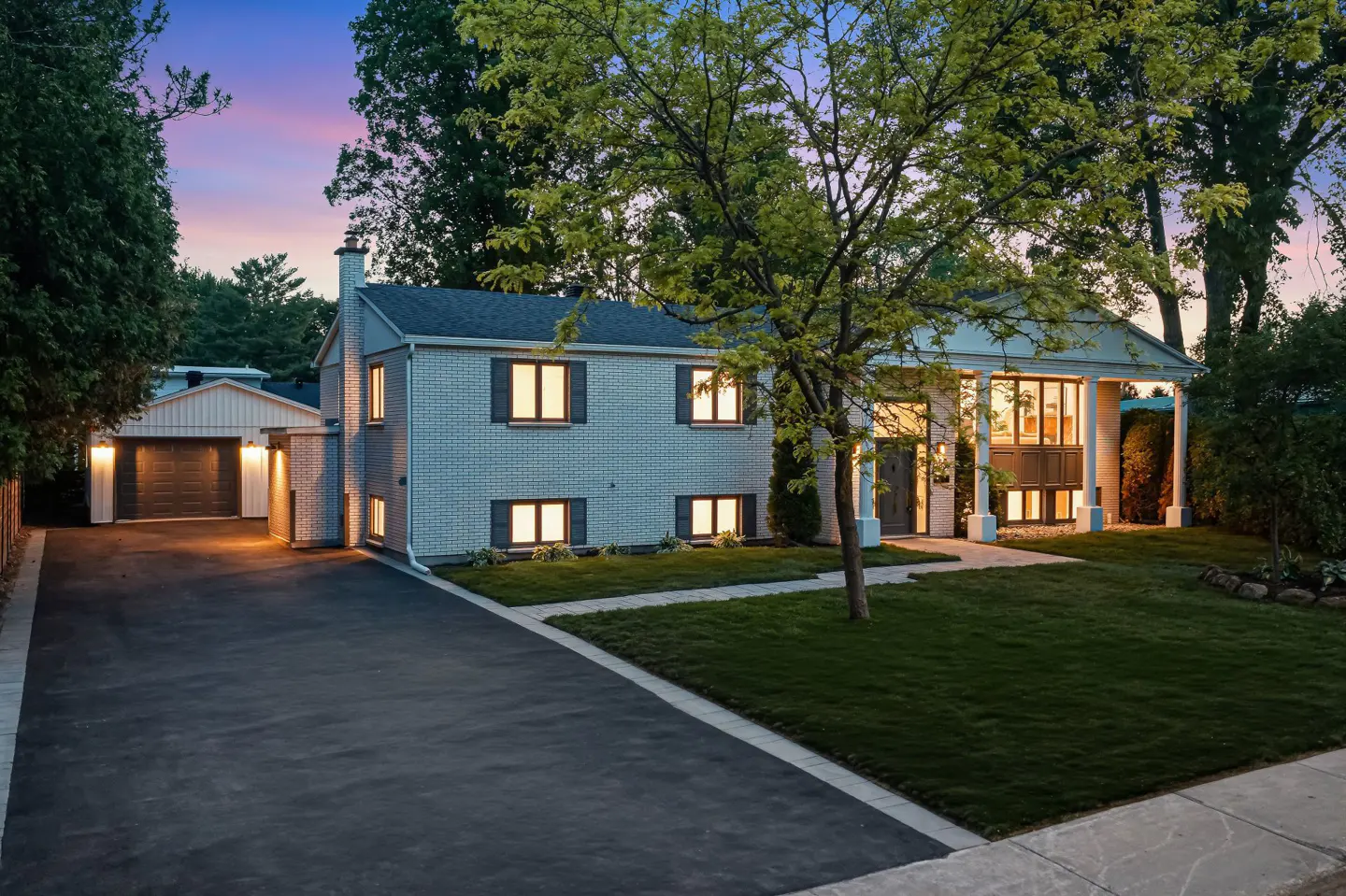 Two-story white brick home at dusk with a paved driveway, detached garage, and illuminated windows under a sunset sky.