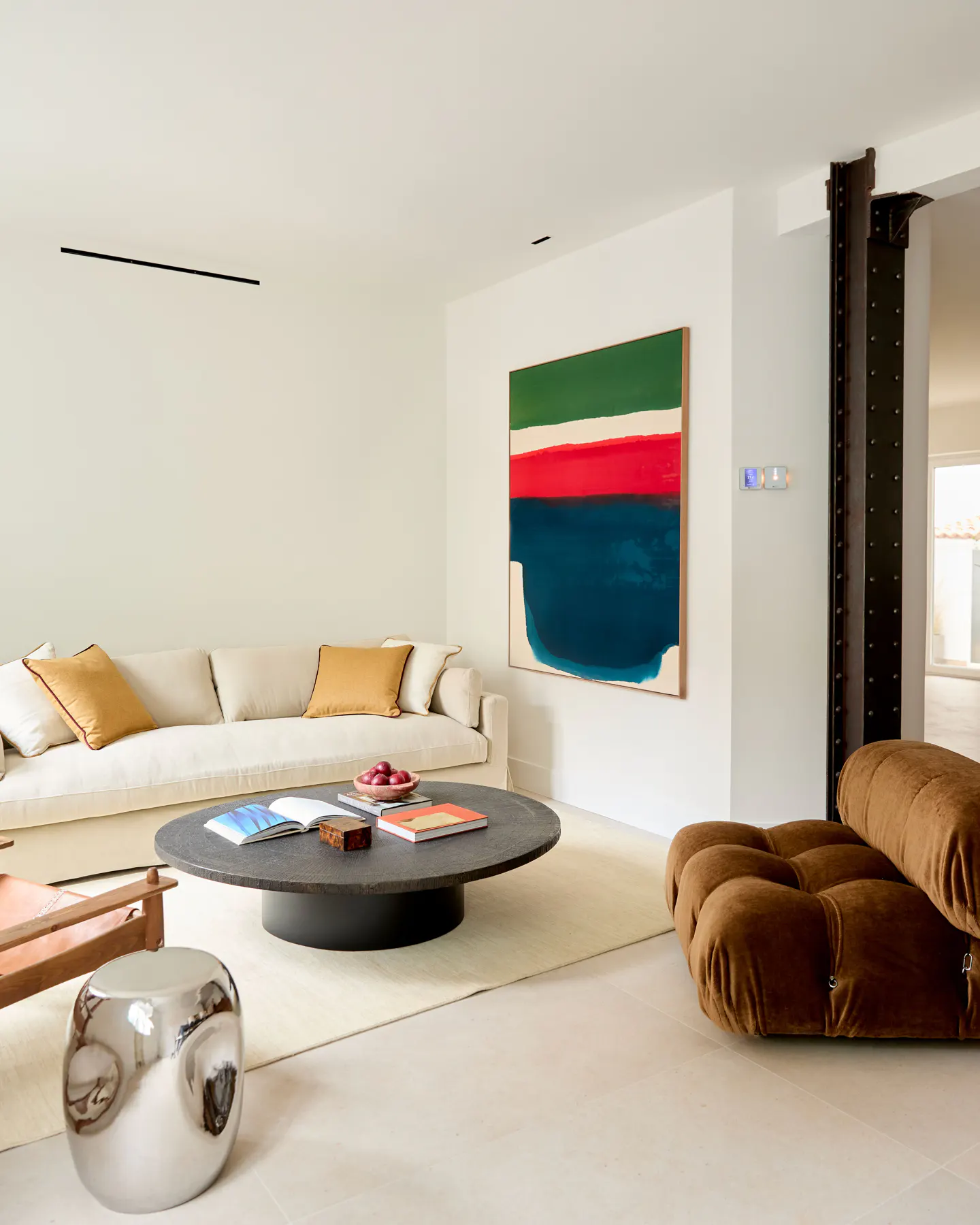 Bright living room with a cream sofa, gold pillows, and a round black coffee table with books and fruit. An abstract painting hangs on the wall.