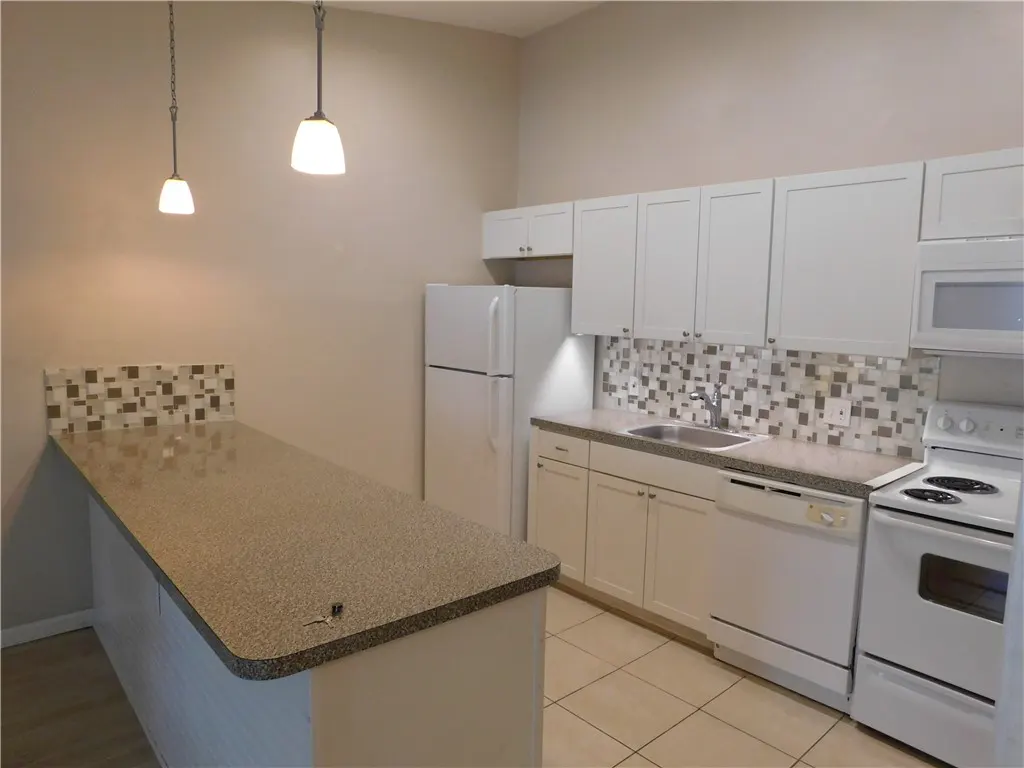 A bright kitchen with white cabinets, appliances, and a speckled countertop island. Two pendant lights hang above the island.