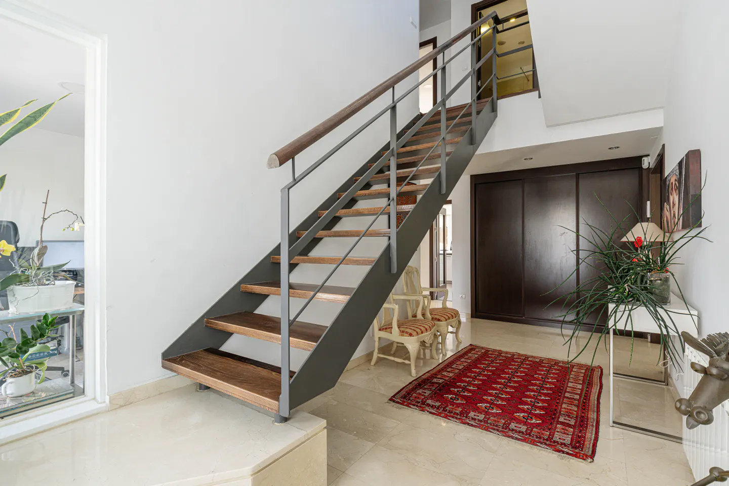 Interior view of a home featuring a staircase with wood steps and metal railings, a red rug, and white walls.