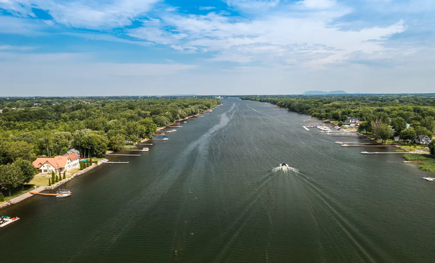 Aerial view of a wide, dark river with green, tree-lined banks under a blue, cloudy sky. A boat moves upriver, leaving a white wake. Houses and docks dot the shoreline.