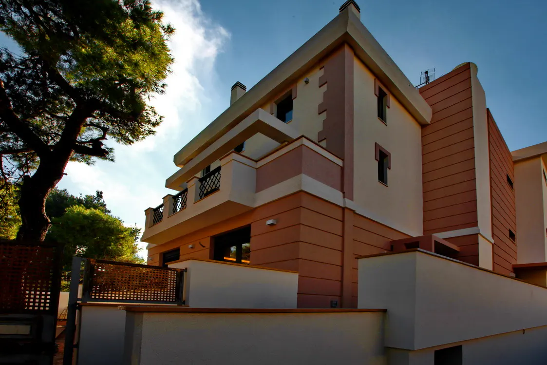 Exterior view of a three-story building with a balcony, painted in peach and white, under a blue sky with a tree on the left.