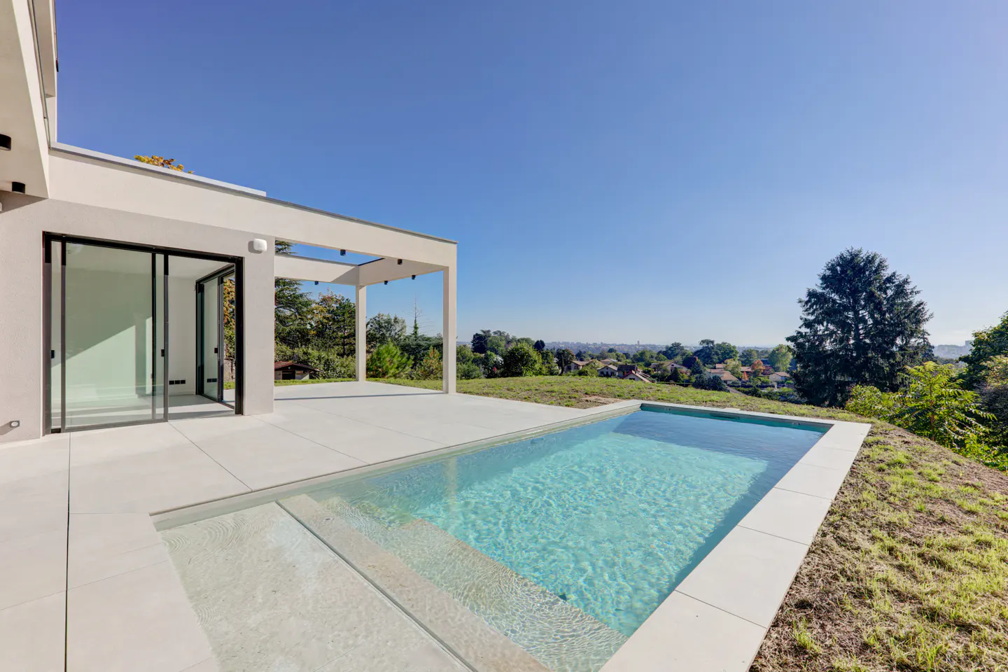Modern home exterior with a rectangular pool, white patio, and sliding glass doors under a clear blue sky.