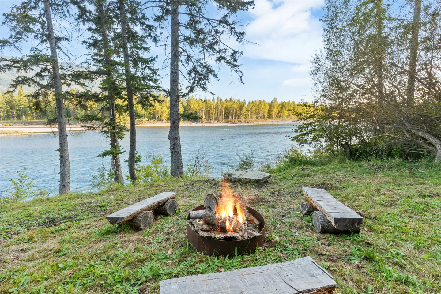 Outdoor fire pit with log benches overlooks a wide river and forest under a blue sky.