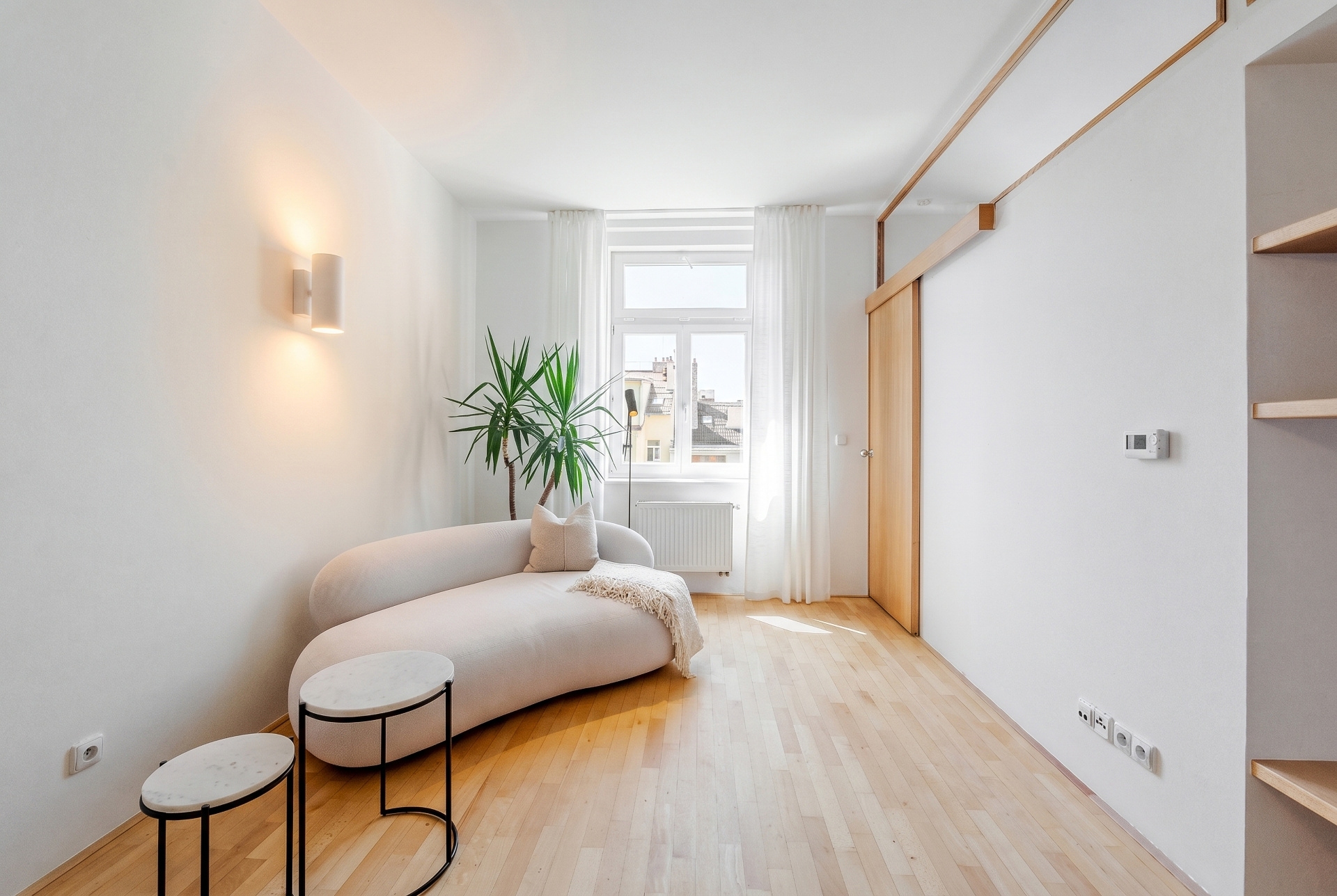 Bright, modern living room with a curved white sofa, two marble-topped tables, and a large window with sheer curtains. A potted plant sits beside the sofa.