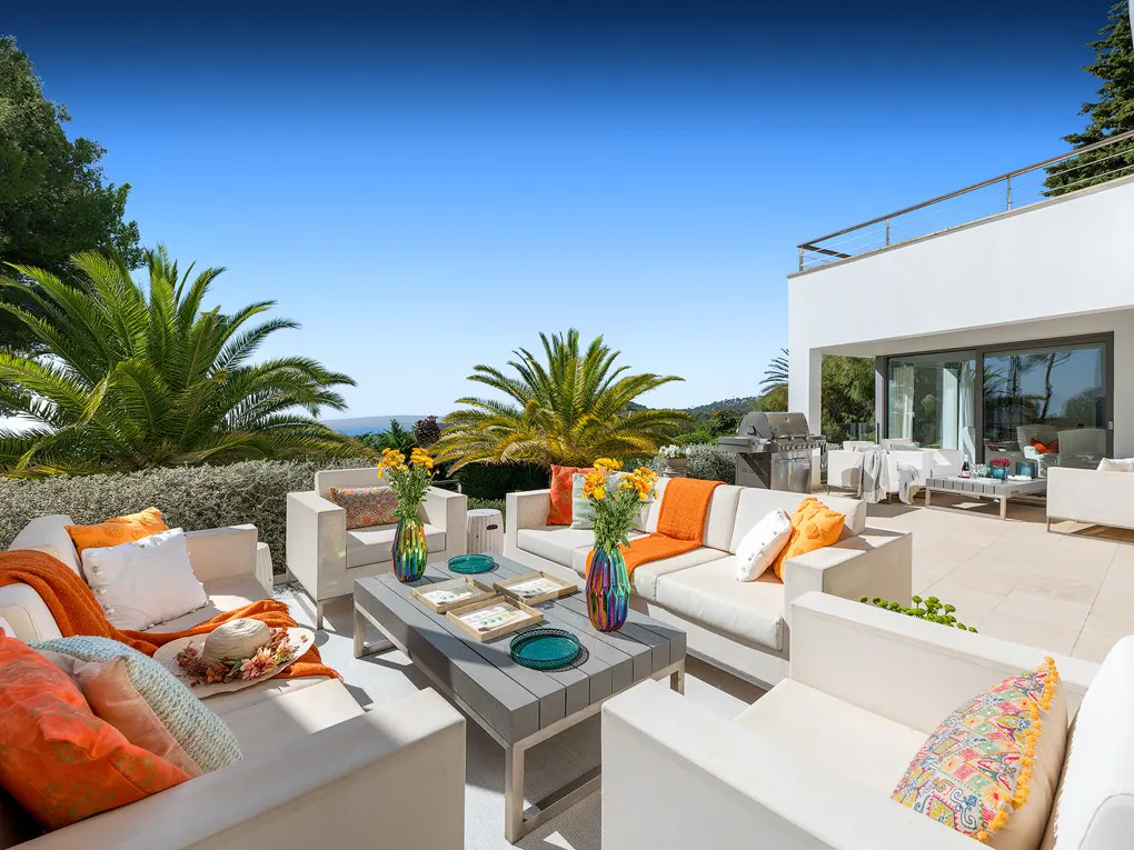Outdoor patio with white sofas, orange pillows, and a gray coffee table with flowers. Palm trees and blue sky in the background.