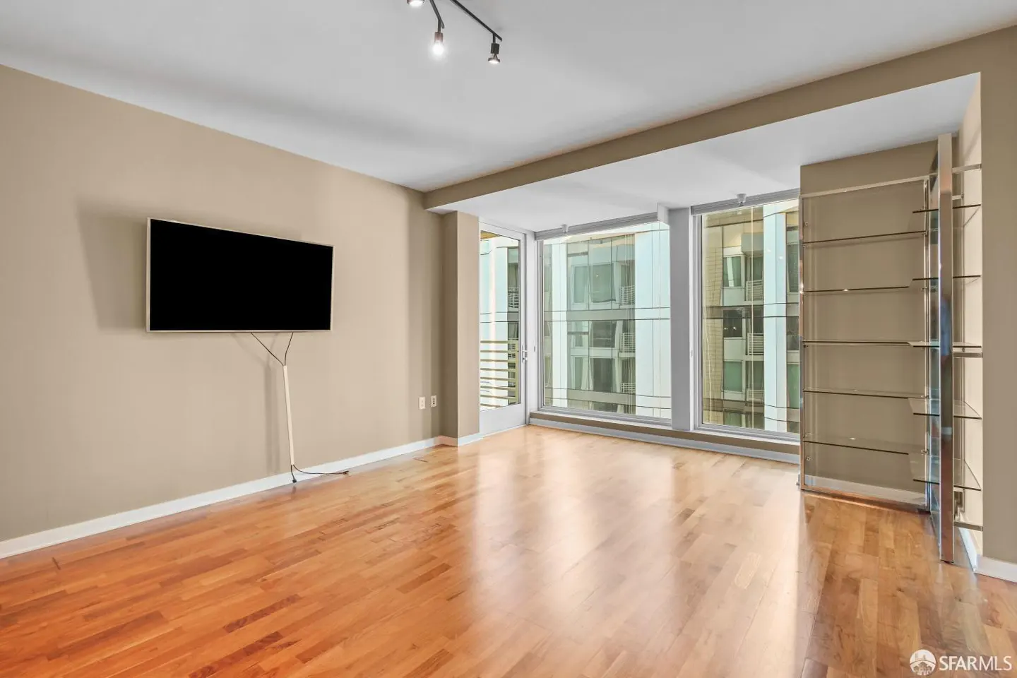 Bright, empty living room with hardwood floors, large windows, and a wall-mounted TV. A chrome shelving unit stands near the windows.