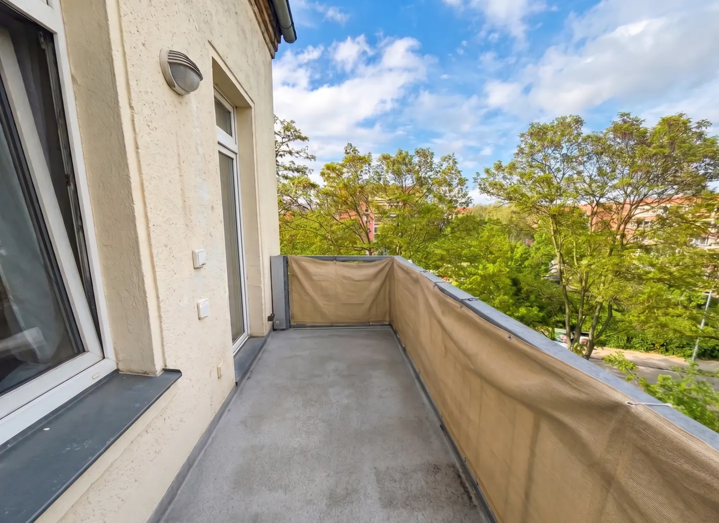 A balcony with a concrete floor and a beige fabric screen, offering a view of green trees and a blue sky.