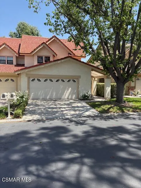 A tan townhouse with a red tile roof and a white garage door is seen on a sunny day. A large tree is on the right.
