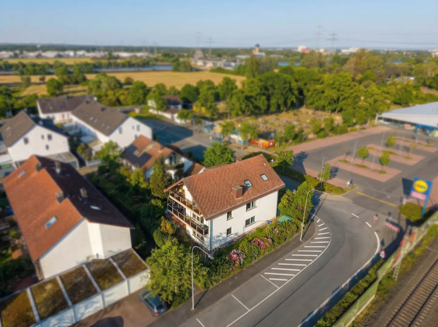 Aerial view of a two-story white house with a red tile roof and a balcony, surrounded by green trees and a road.