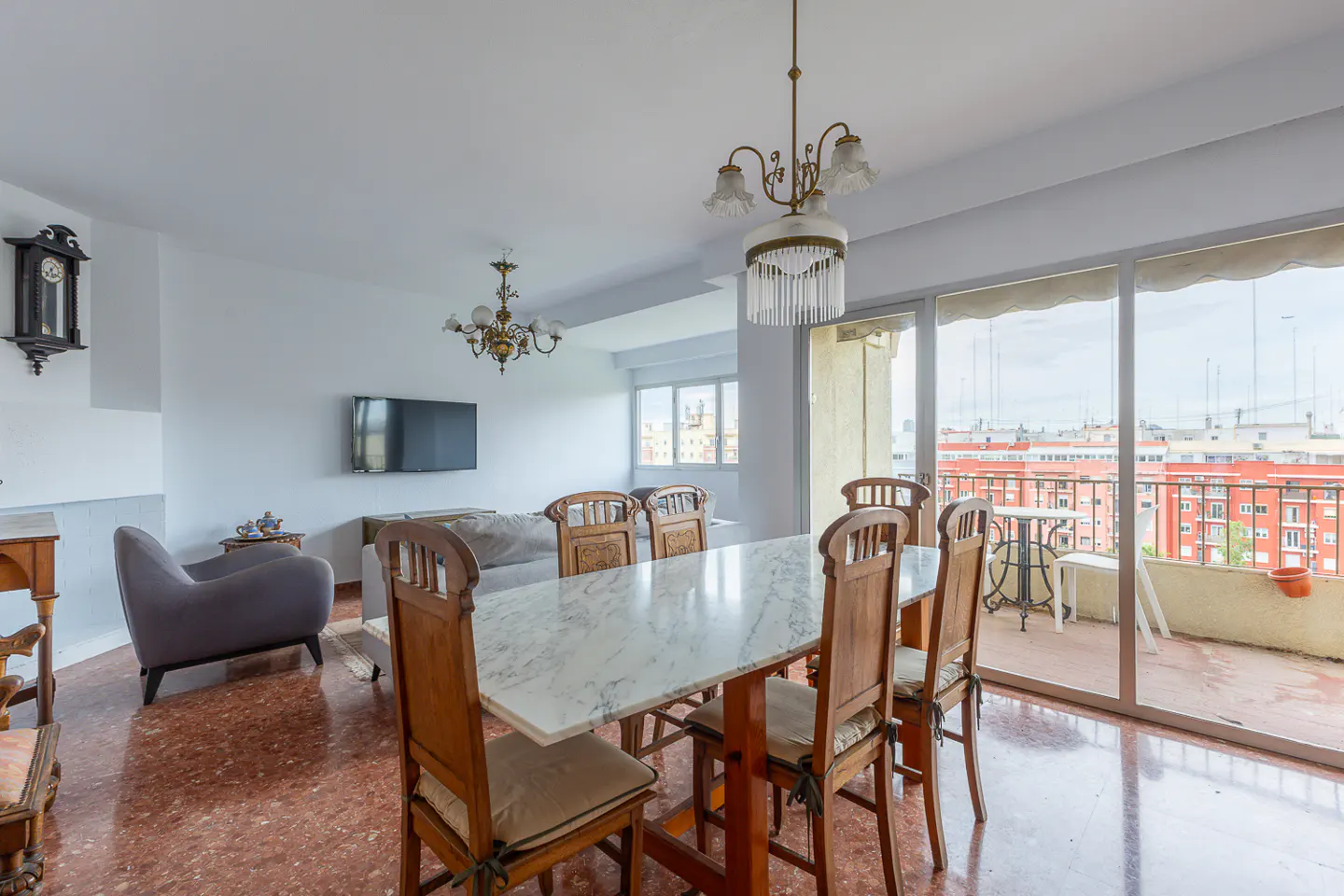Bright living room with marble dining table, wooden chairs, and balcony view of red buildings. Chandeliers hang from the white ceiling.