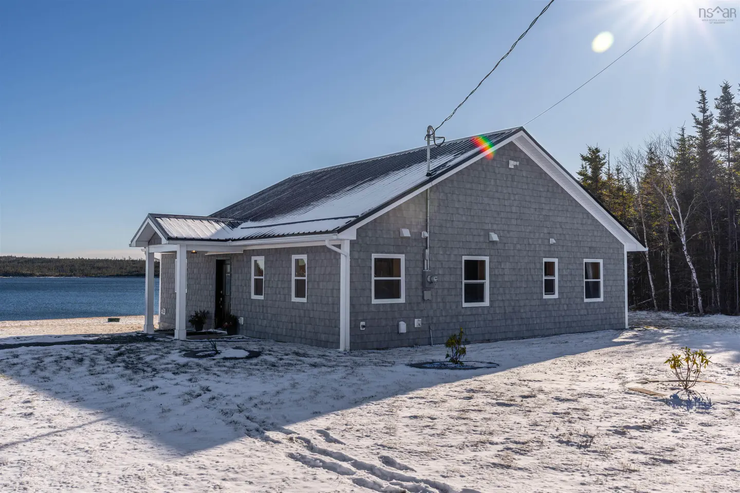 Gray shingle house with a metal roof and white trim on a snowy lawn near a body of water under a clear blue sky.