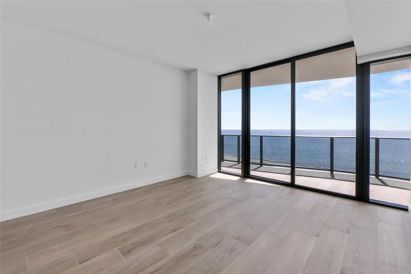 Empty room with light wood floors, white walls, and floor-to-ceiling windows overlooking the ocean.