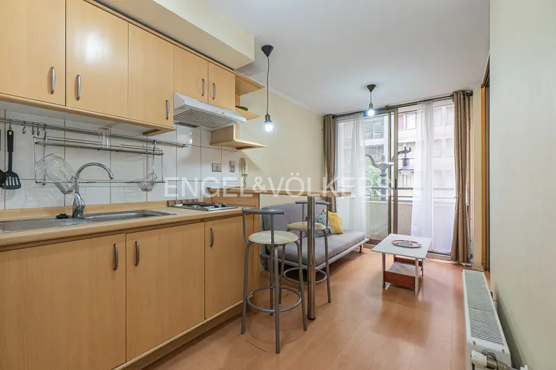 A bright studio apartment with light wood cabinets, a small sofa, and a balcony with sheer curtains.