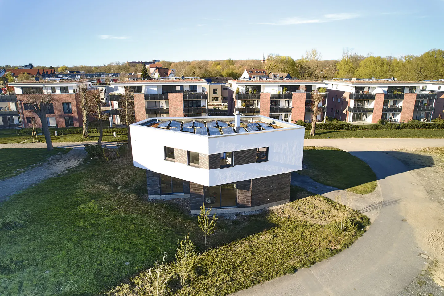 Modern two-story house with white exterior, dark brick base, and solar panels on the roof. Other houses and trees in background.