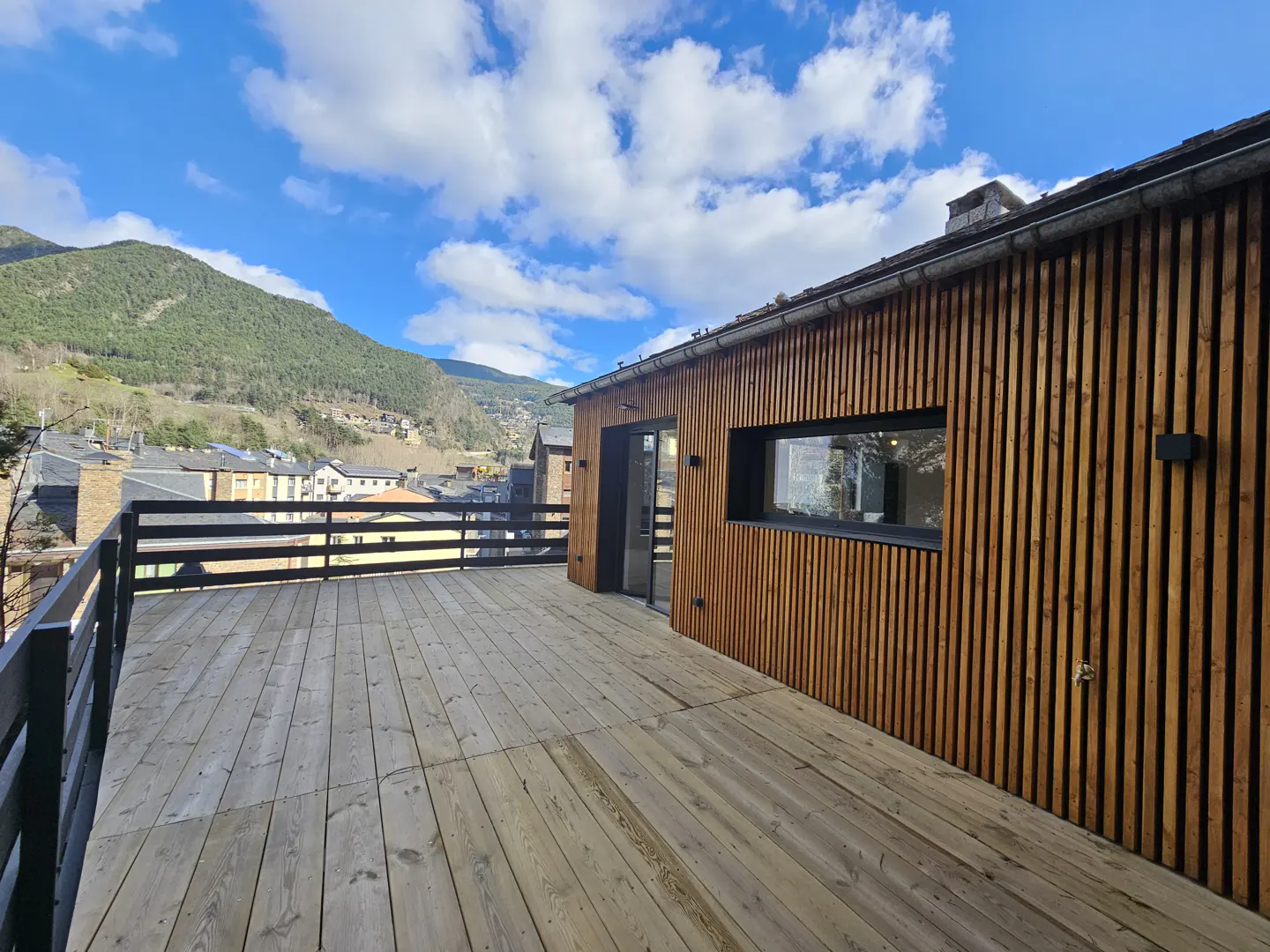 Wide wooden deck with black railing, attached to a wood-paneled building. Mountain view in the background under a blue, cloudy sky.