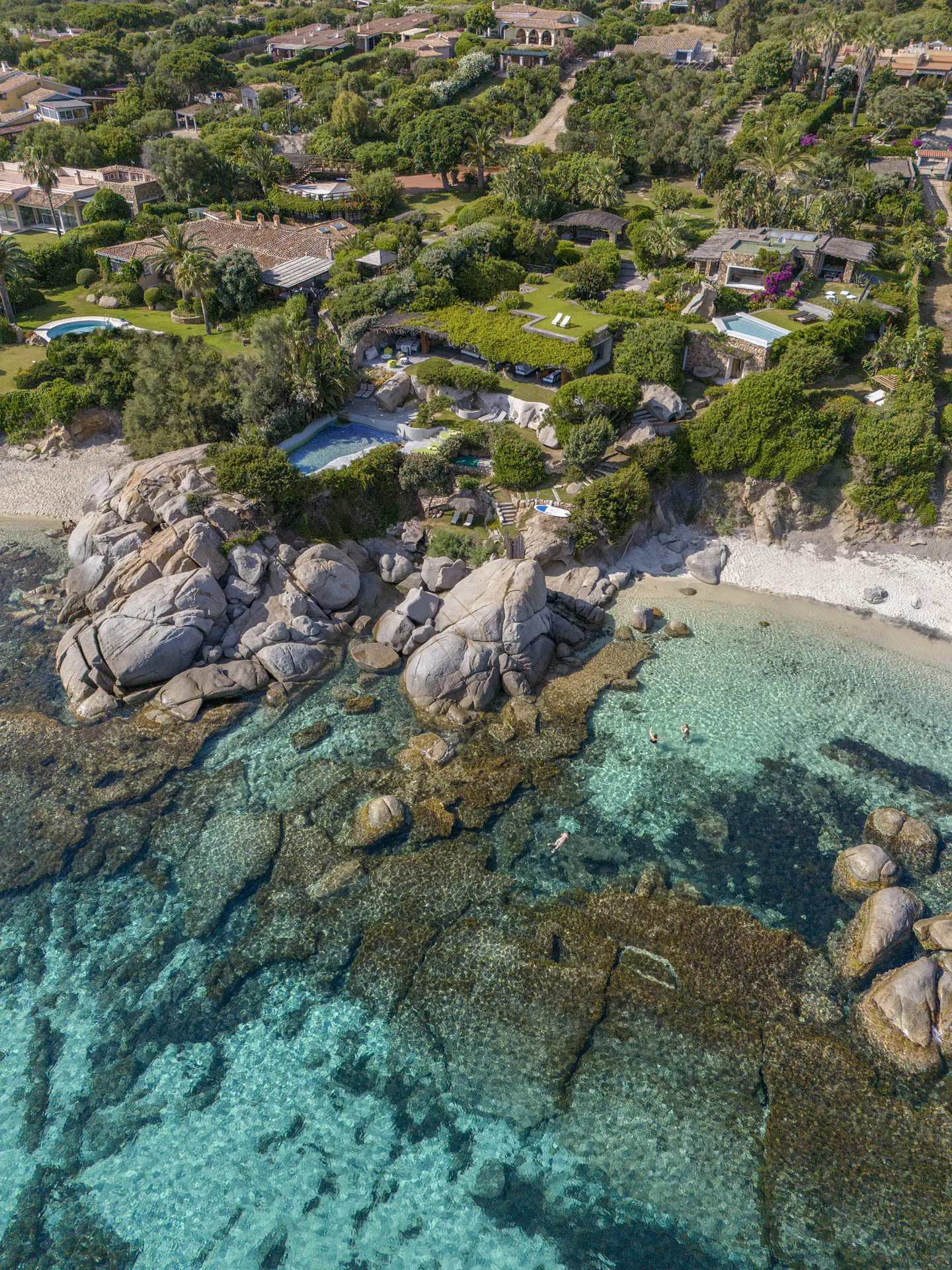 Aerial view of a rocky coastline with turquoise water, villas, and lush green vegetation.