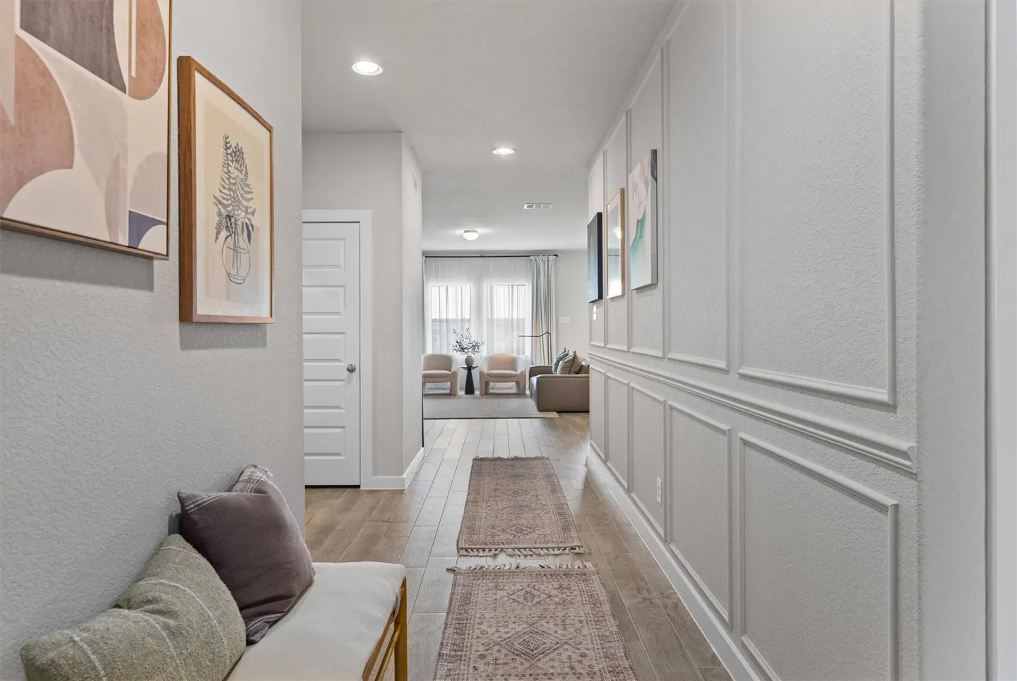 Hallway with light gray walls, wood floors, and a long rug. A white bench with pillows sits near framed art. A living room is visible at the end.