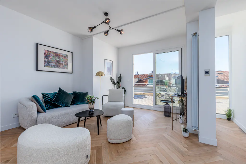 Bright living room with white walls, herringbone wood floors, and a balcony view. A couch with green pillows and ottomans are in the foreground.
