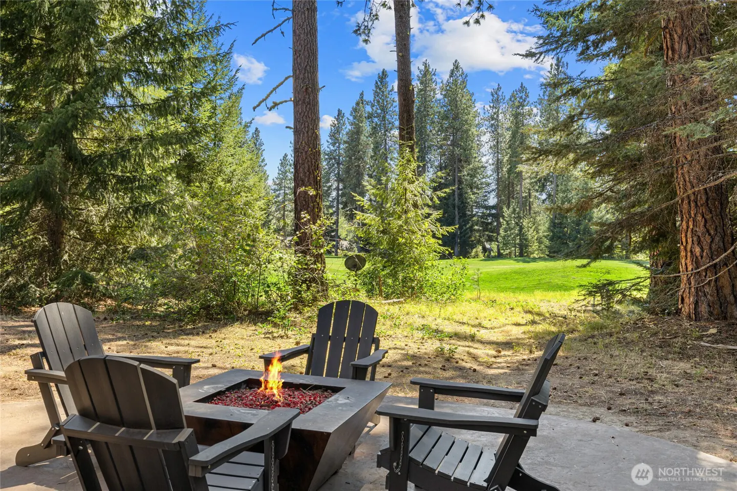 Outdoor patio with four black Adirondack chairs around a square fire pit with flames, overlooking a green lawn and pine trees under a blue sky.