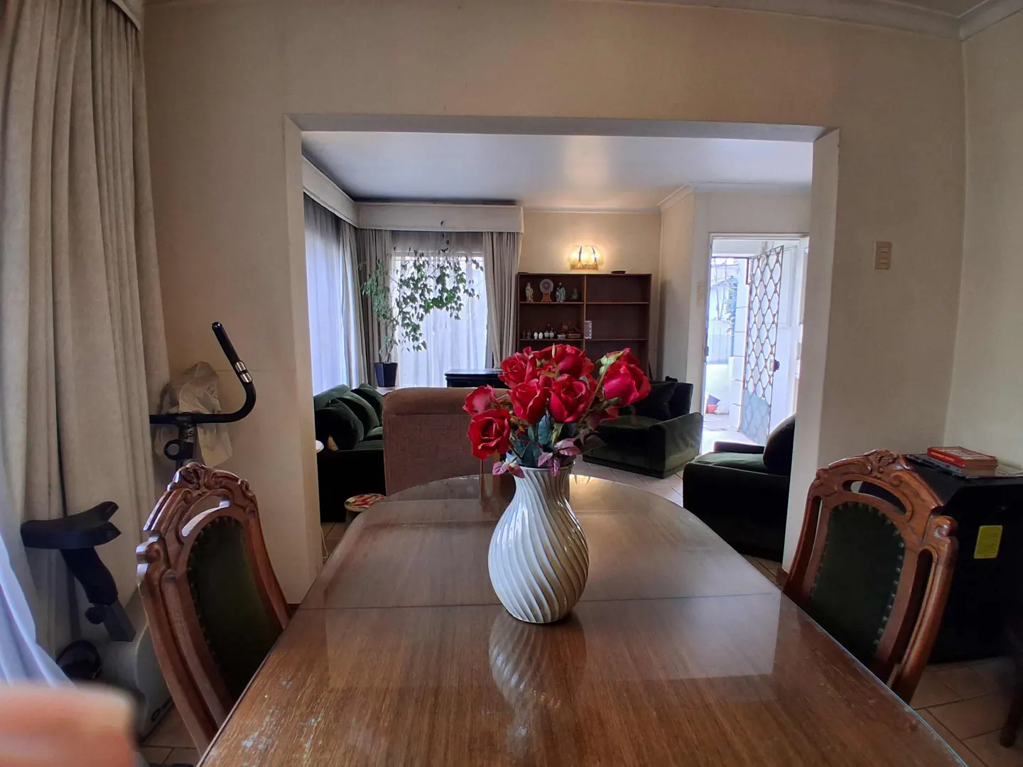 A dining room with a wooden table and chairs, a white vase with red roses, and a view into the living room with green sofas.