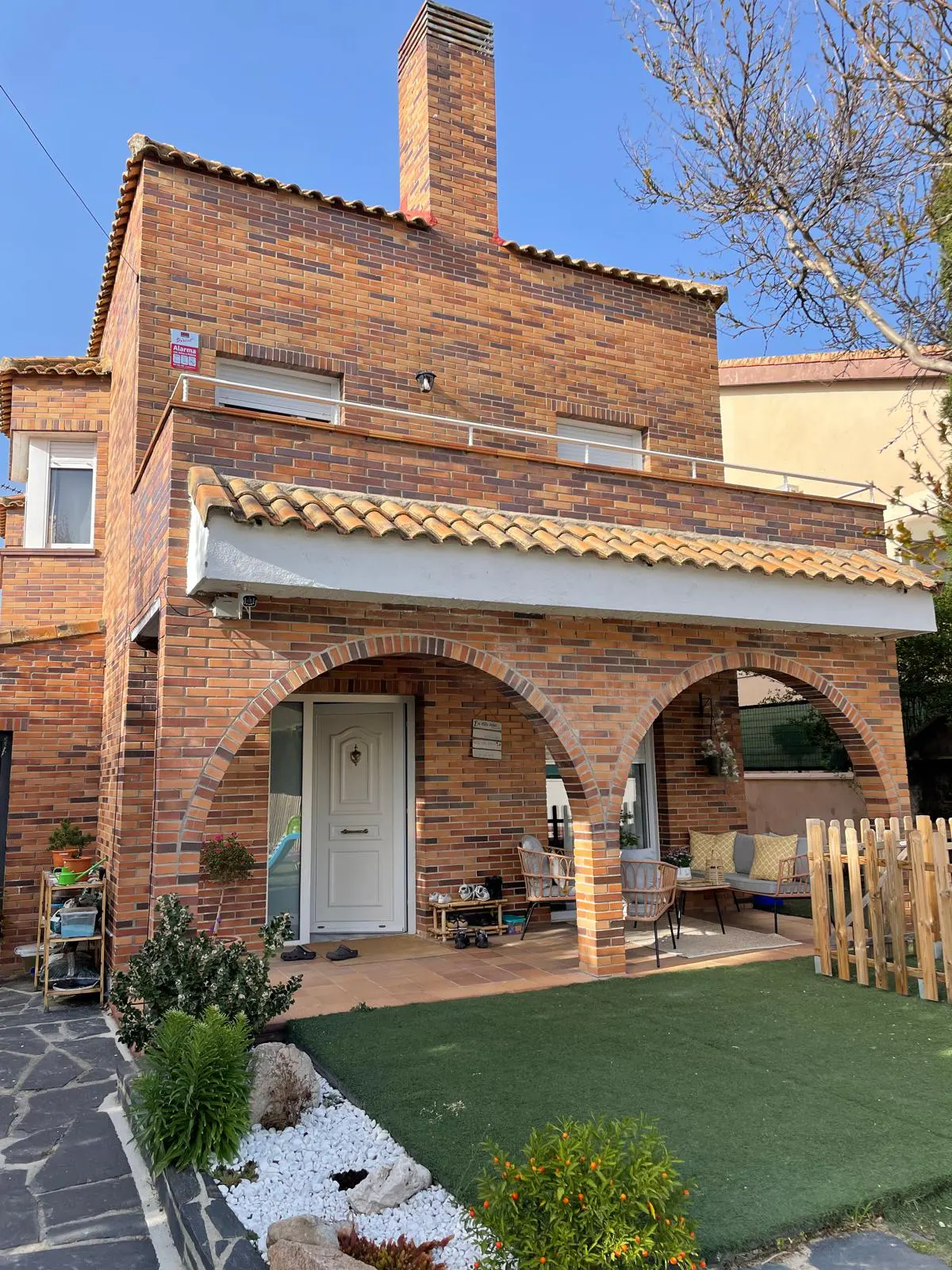 Two-story brick house with arched porch, white door, and chimney. Green lawn and stone walkway lead to the entrance.