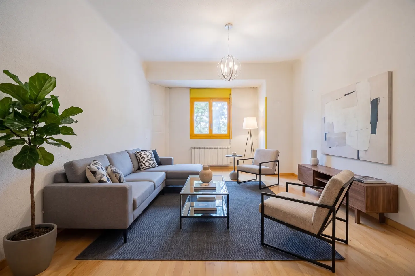 Bright living room with a gray sectional sofa, two chairs, a glass coffee table, and a large potted plant. A yellow-framed window adds a pop of color.