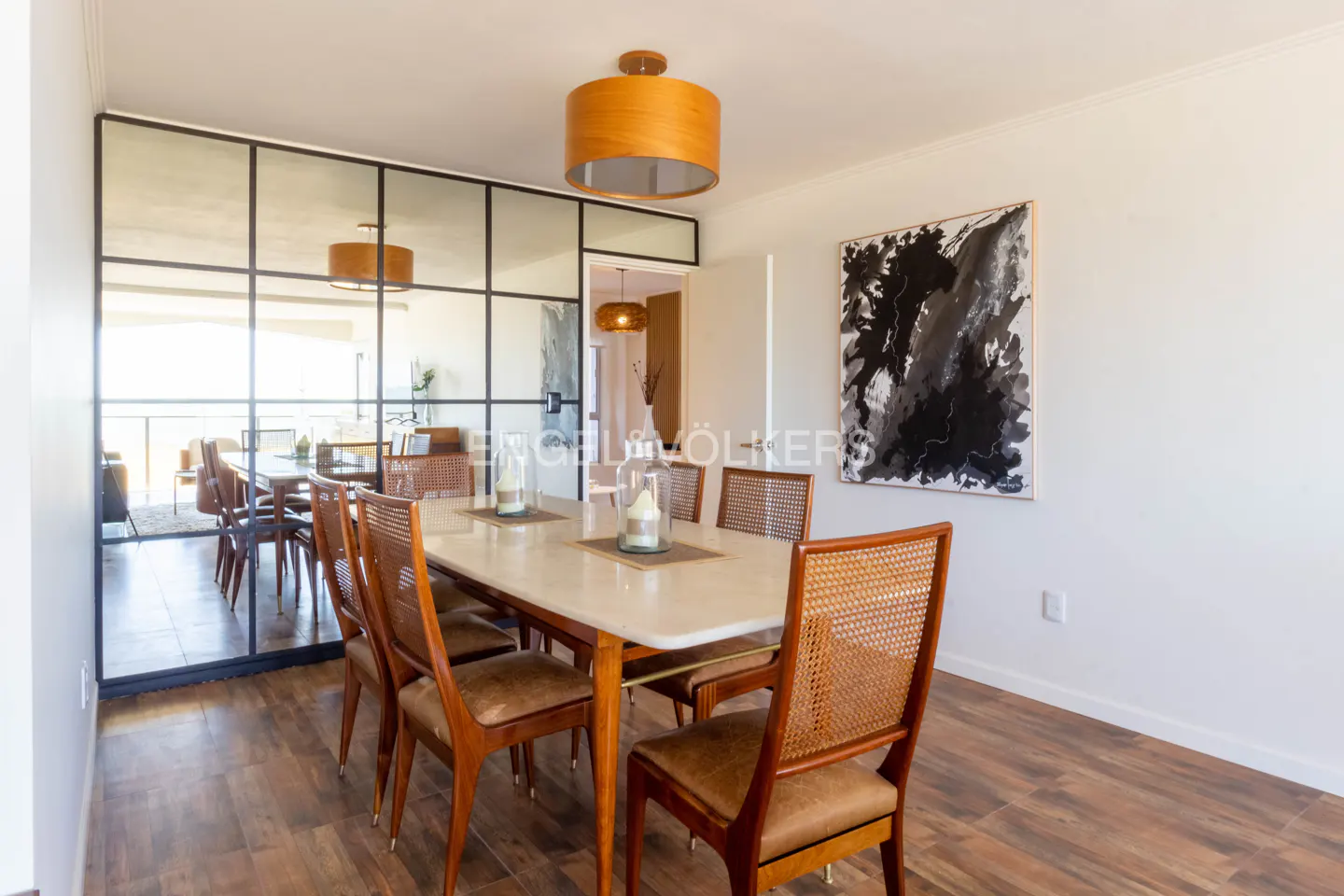 A dining room with a white table, wooden chairs, and a black and white painting on the wall. A glass partition separates the dining area from another room.