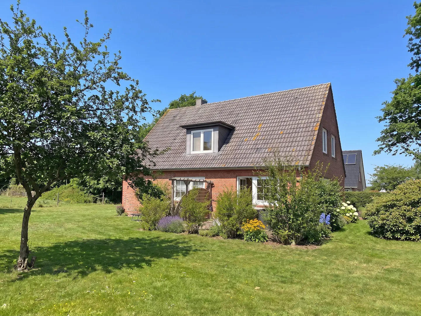 A red brick house with a gray tiled roof sits on a green lawn under a blue sky. A tree is on the left.