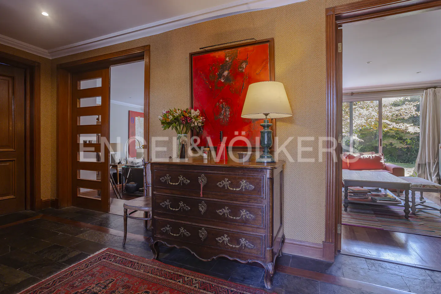 Entryway with a dark wood dresser, lamp, and flowers. A red painting hangs above. A doorway leads to a living room with a red couch.