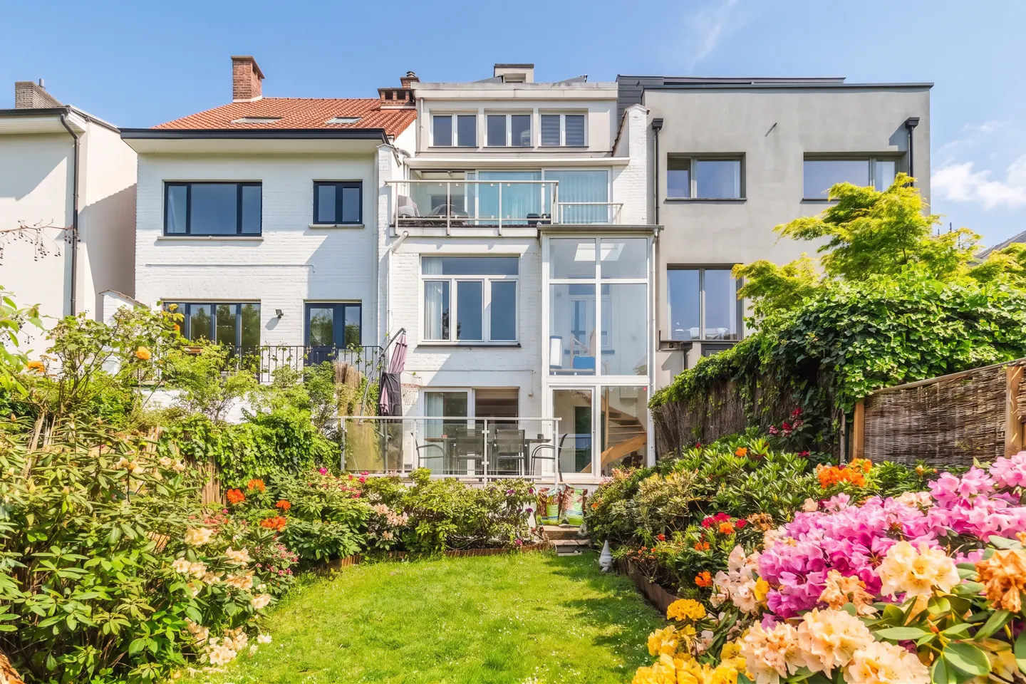 Backyard view of a three-story house with white and gray walls, balconies, and a garden with colorful flowers.