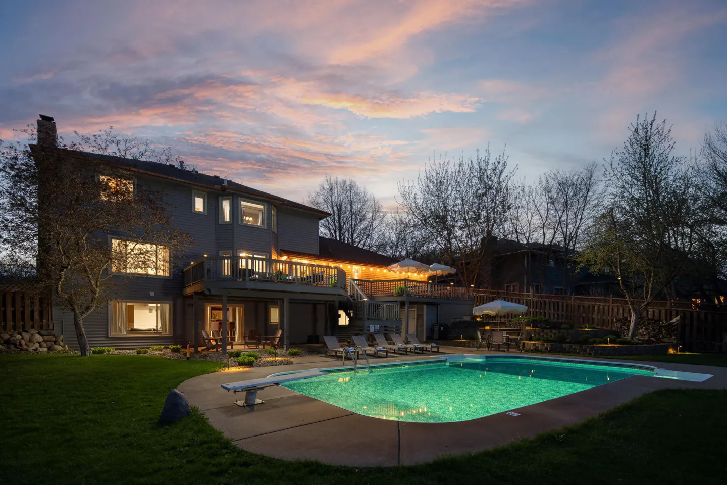 Backyard view of a two-story gray house with a pool, deck, and lawn at dusk.