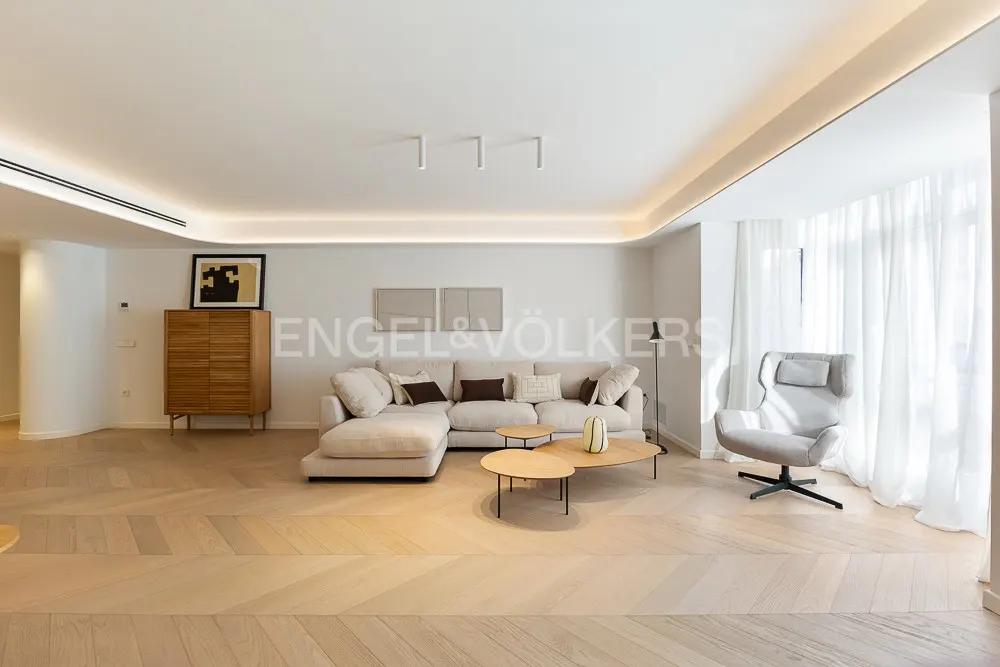 Bright living room with a beige sectional sofa, wooden coffee tables, a gray armchair, and light wood floors. White walls and curtains.