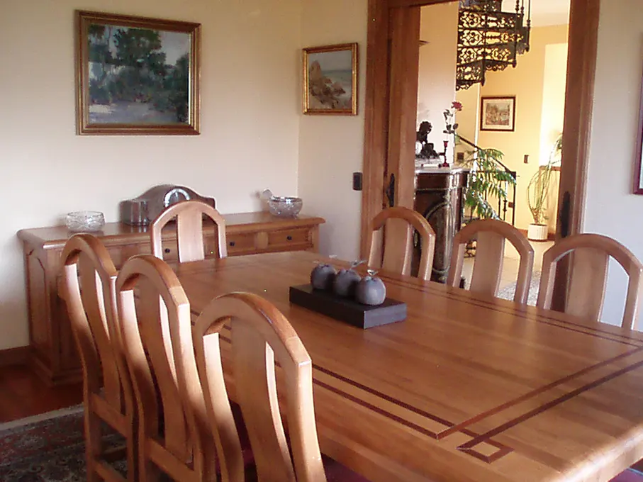 A dining room with a long wooden table and chairs, a buffet, and artwork on the walls. A spiral staircase is visible through an open doorway.