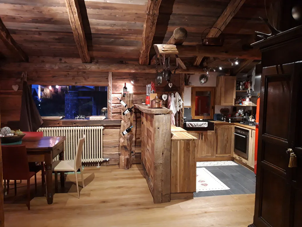 Rustic cabin interior with wood beams, floors, and walls. A dining table sits near a window, and a kitchen with wooden cabinets is visible.